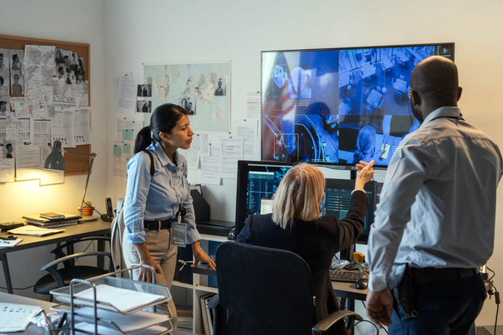 Three law enforcement professionals analyze surveillance footage on multiple monitors in a cluttered security office, with crime-related documents pinned to the walls.