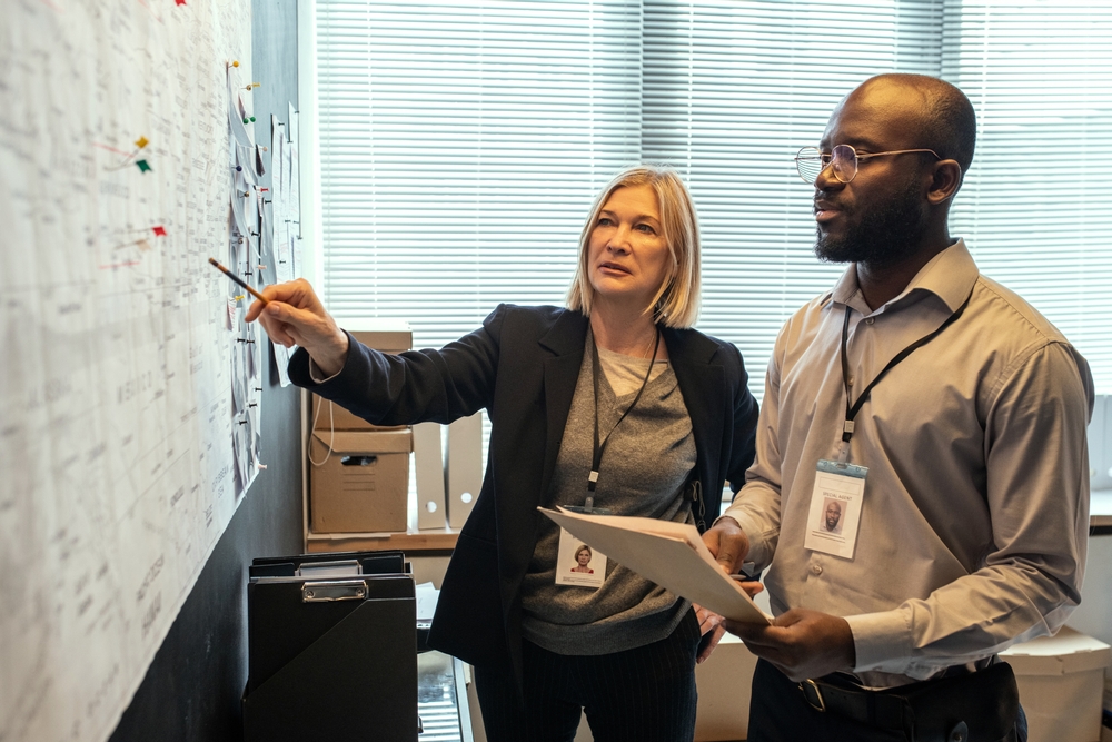 Two professionals discuss a case in an office, with one pointing at a detailed planning board.