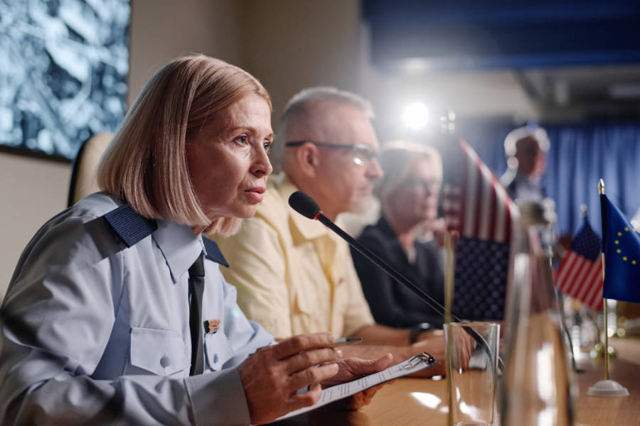 A uniformed woman speaks into a microphone at a conference table. Other panelists sit beside her, with U.S. and European Union flags visible on the table.