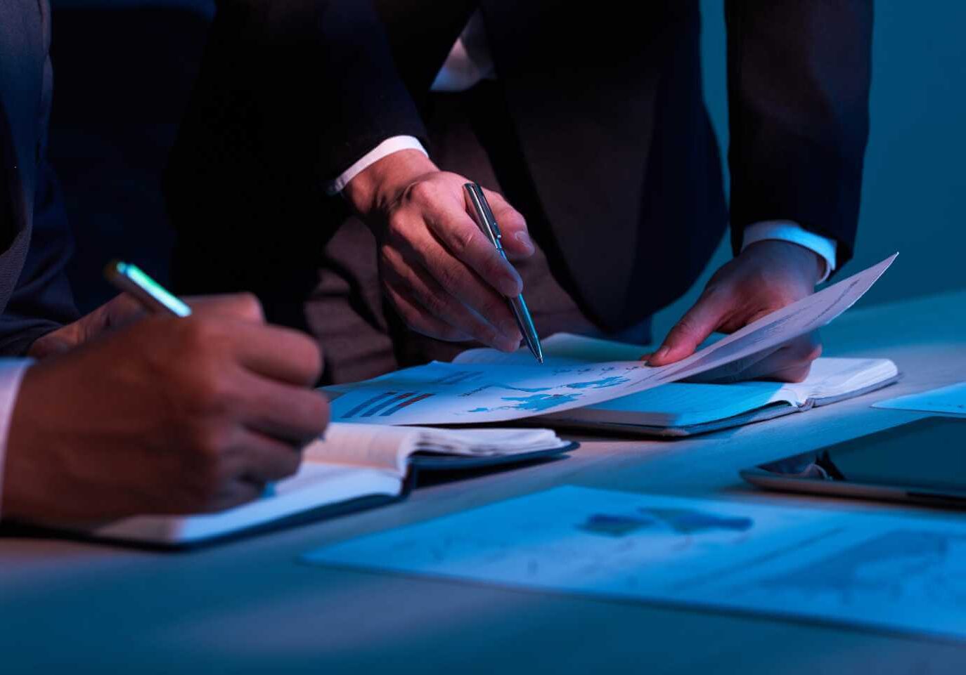 Close-up of two people in suits working at a table, writing notes and reviewing charts on paper documents under low lighting.