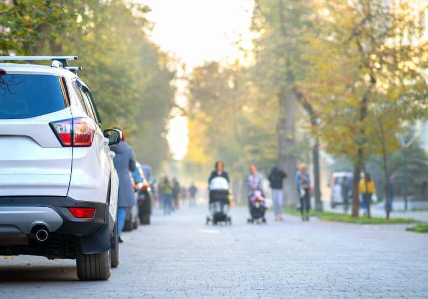 A white SUV is parked on the side of a tree-lined city street. People walk in the background, some pushing strollers, on a bright autumn day.