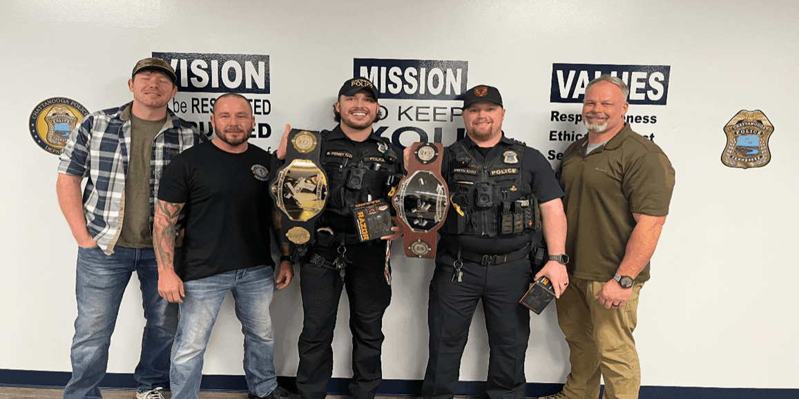 Five men standing in front of a wall with “Vision, Mission, Values” signs. Two officers in uniform hold championship-style belts as awards, while three others in casual clothing stand beside them.