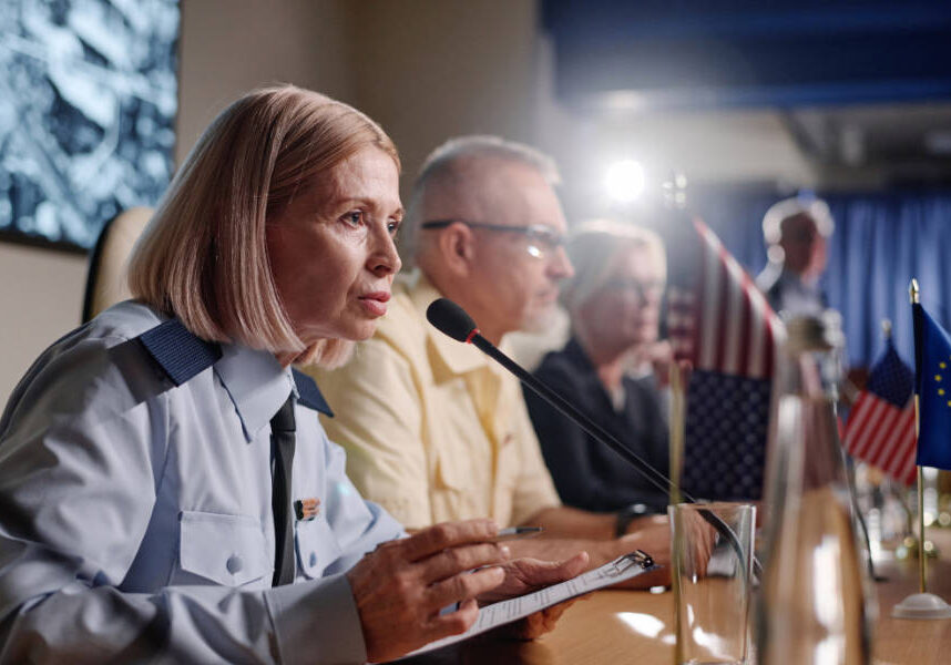 A uniformed woman speaks into a microphone at a conference table. Other panelists sit beside her, with U.S. and European Union flags visible on the table.