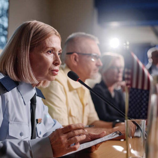A uniformed woman speaks into a microphone at a conference table. Other panelists sit beside her, with U.S. and European Union flags visible on the table.