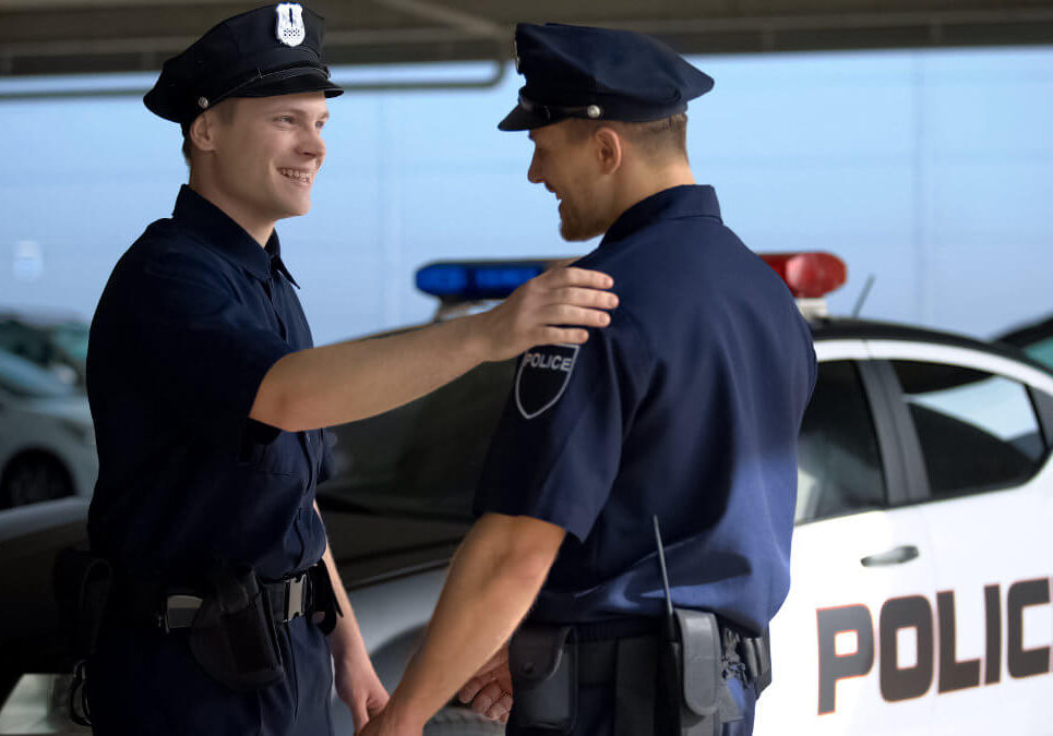 Two police officers in uniform smiling and greeting each other beside a patrol car.